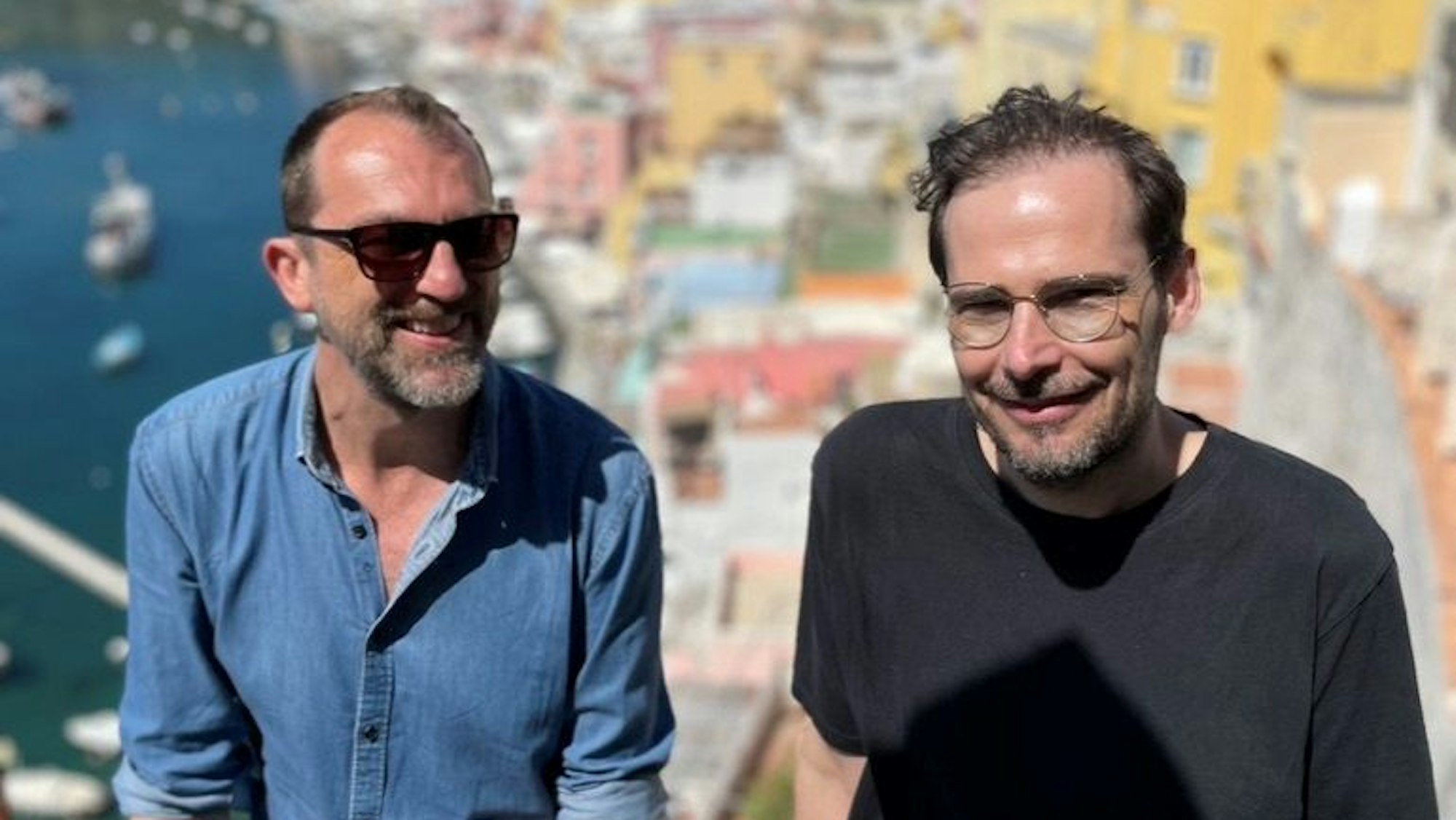 Michael Dietz (links) und Jochen Schliemann sitzen lächeln nebeneinander, im Hintergrund eine sonnenbeschienene, bunte Stadt.