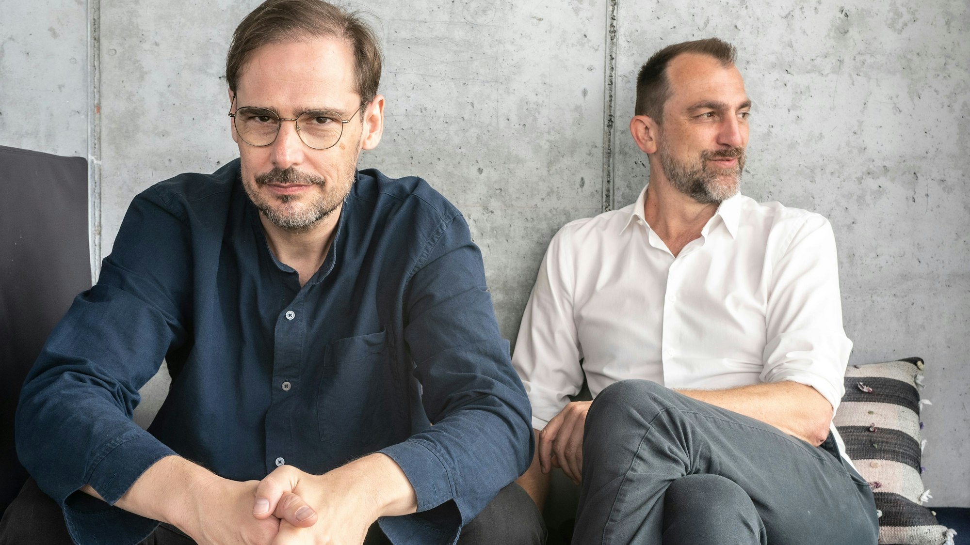 Die Podcaster und Autoren Jochen Schliemann (links) und Michael Dietz sitzen nebeneinander vor einer grauen Wand.