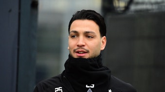 Ramy Bensebaini von Borussia Mönchengladbach, hier bei einer Trainingseinheit am 31. Januar 2023 in schwarze Kleidung und Schal eingepackt. Er lächelt bein Betreten des Platzes leicht.