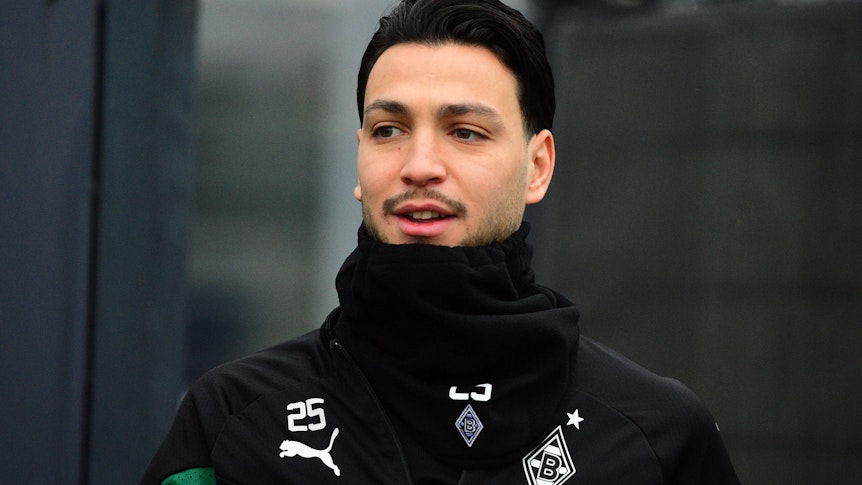 Ramy Bensebaini von Borussia Mönchengladbach, hier bei einer Trainingseinheit am 31. Januar 2023 in schwarze Kleidung und Schal eingepackt. Er lächelt bein Betreten des Platzes leicht.