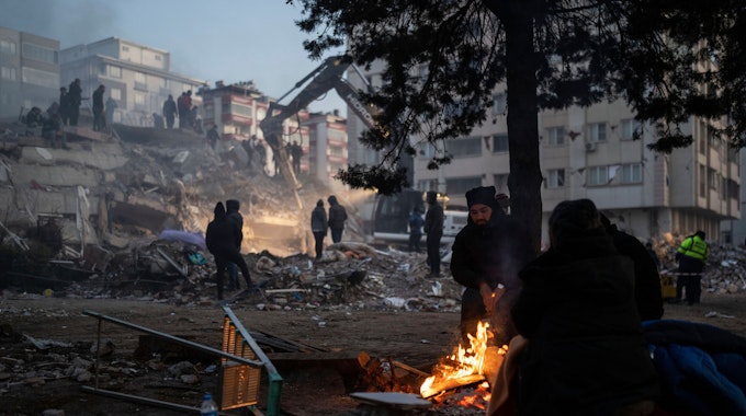 Türkei, Kahramanmaras: Menschen wärmen sich an einem Feuer, während Retter ein durch die Erdbeben zerstörtes Gebäude durchsuchen.