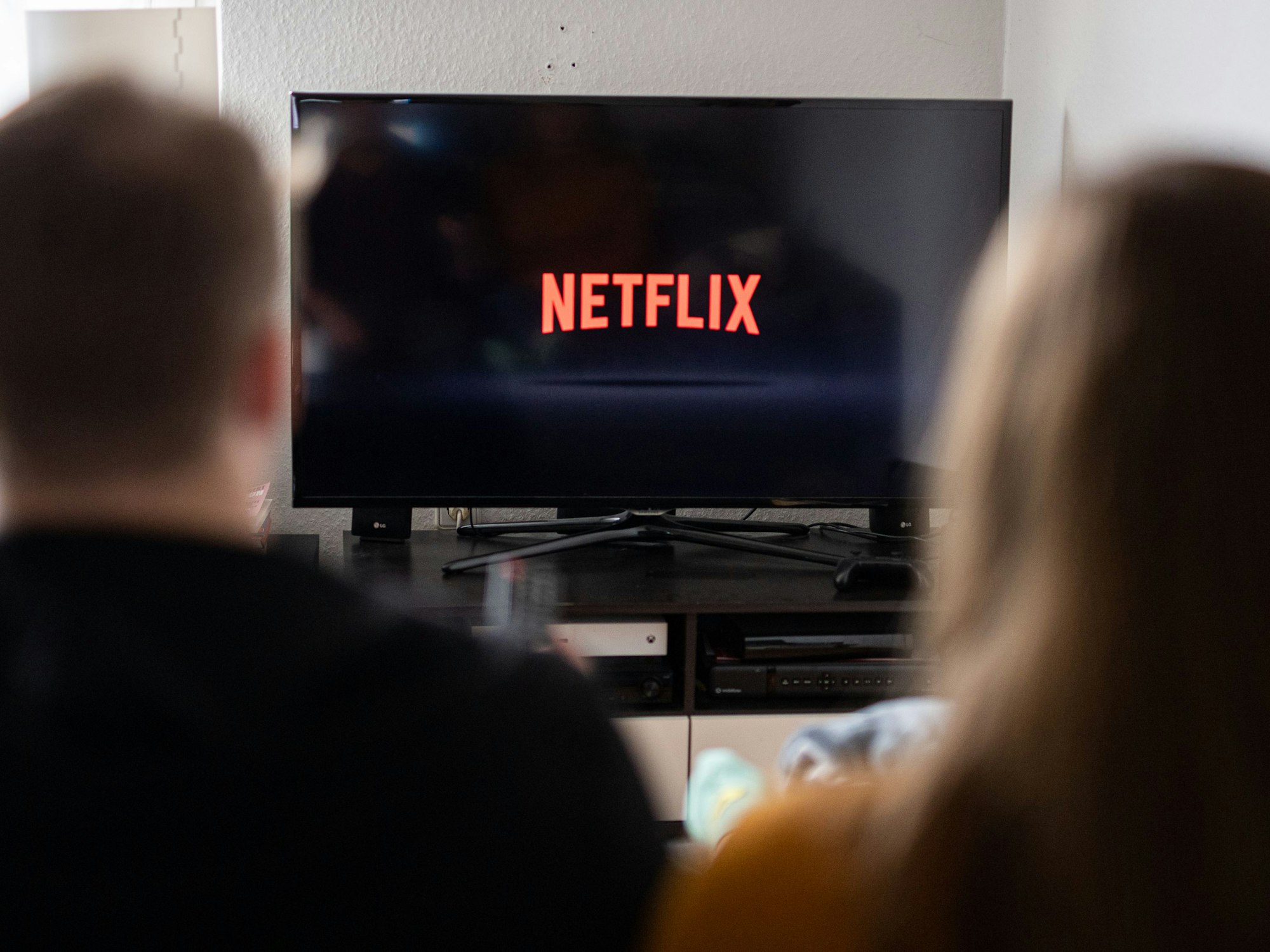 Zwei Personen sitzen vor einem Fernseher, auf welchem das Logo des Streaming-Anbieters Netflix zu sehen ist.