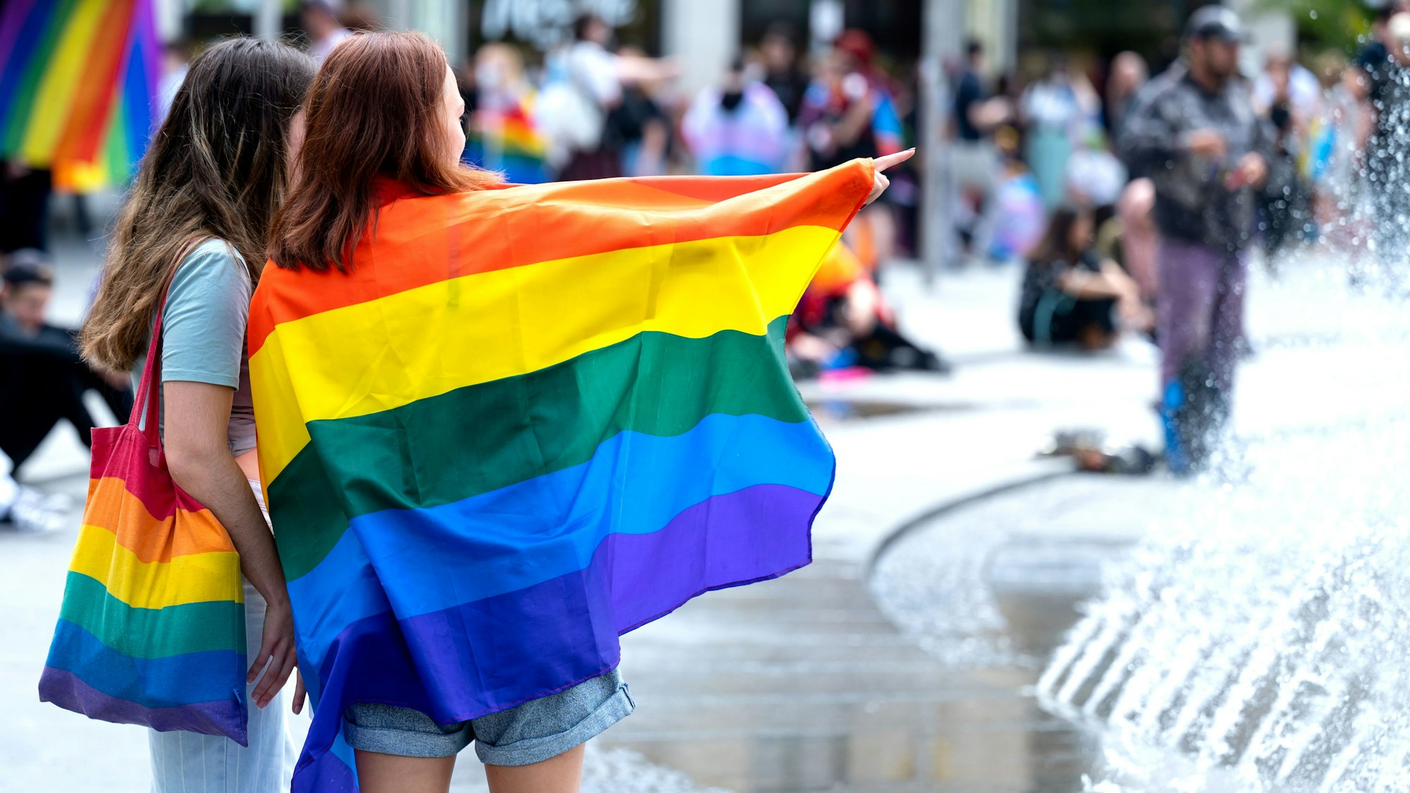 Das undatierte Symbolfoto zeigt zwei Frauen mit Regenbogentasche bzw. einer umgehängten Fahne in Regenbogenfarben. Hinter ihnen befinden sich weitere Menschen.