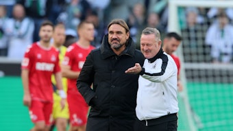 Daniel Farke (l.) und Roland Virkus stehen nach dem Bundesliga-Sieg im Derby gegen den 1. FC Köln auf dem Rasen im Borussia-Park nebeneinander und blicken in Richtung der Nordkurve. Virkus hebt seinen linken Arm und zeigt nach vorne.