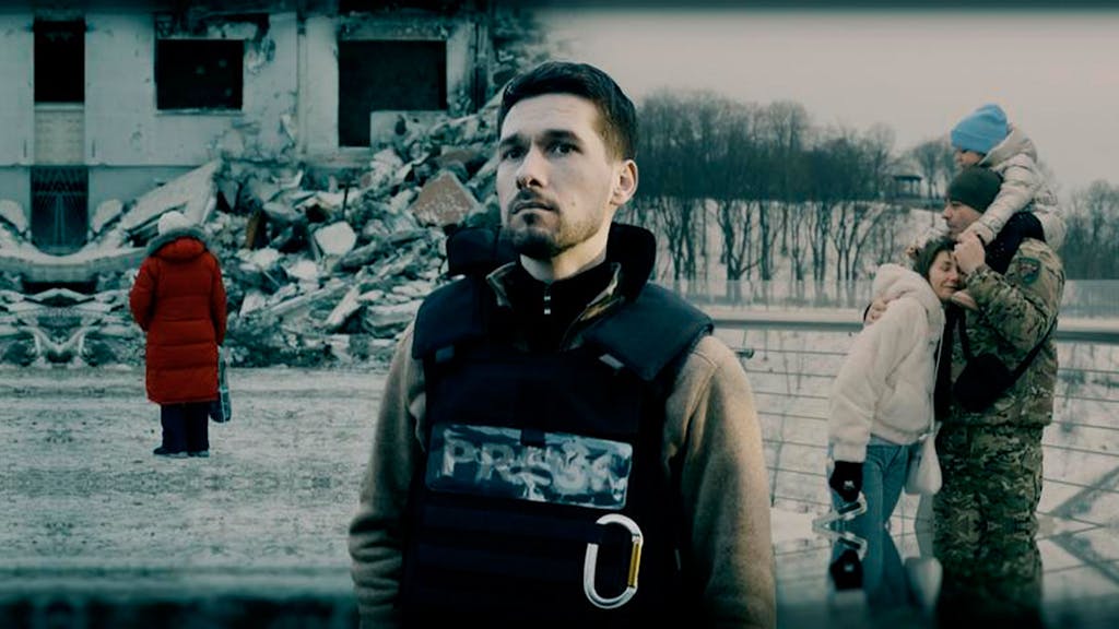 Das Foto zeigt eine Szene aus dem ARD-Film „Ukraine – Krieg im Leben“. Ein Mann mit Schutzweste steht im Vordergrund, hinter ihm sind eine junge Familie und ein zerstörtes Gebäude zu sehen.