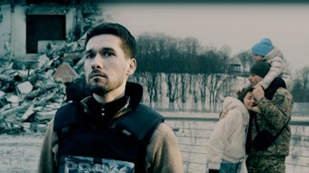 Das Foto zeigt eine Szene aus dem ARD-Film „Ukraine – Krieg im Leben“. Ein Mann mit Schutzweste steht im Vordergrund, hinter ihm sind eine junge Familie und ein zerstörtes Gebäude zu sehen.