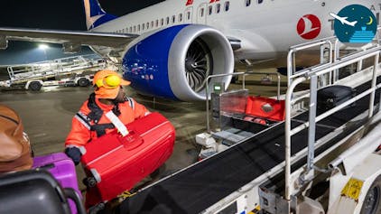 Ein Mitarbeiter der Gepäckabfertigung wuchtet einen roten Koffer auf ein Förderband. Im Hintergrund ist ein Flugzeug zu sehen.&nbsp;