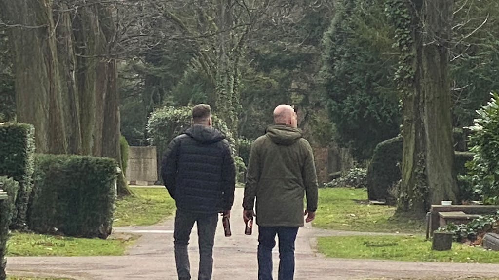 Zwei Männer gehen mit Bierflaschen in der Hand über den Friedhof Melaten.