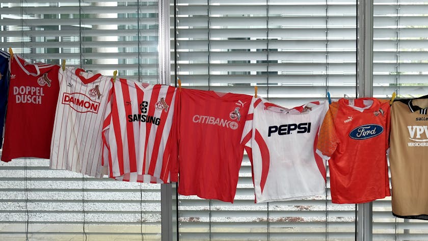 Verschiedenfarbige Trikots des 1. FC Köln hängen auf einer Wäscheleine. Sie zeigen die verschiedenen Sponsoren-Logos der Vereinsgeschichte.