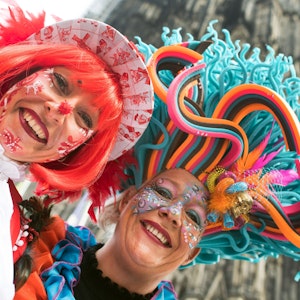 Das Symbolfoto zeigt zwei zu Weiberfastnacht verkleidete Frauen mit bunten Hüten und Karnevalsschminke im Gesicht. Im Hintergrund steht der Kölner Dom.