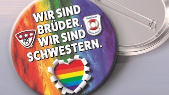 Der Pin ist in Regenbogen-Farben gestaltet, darauf steht ein Zitat aus dem Brings-Song „Liebe gewinnt“: „Wir sind Brüder, wir sind Schwestern“.