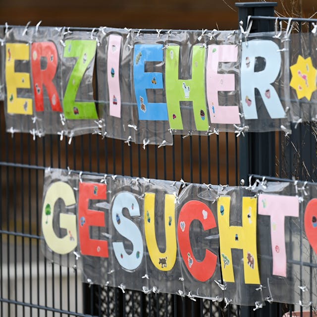 Mit bunten Buchstaben sind die Worte "Erzieher*in gesucht" geschrieben, die an einem Zaun einer Kindertagesstätte befestigt sind.&nbsp;