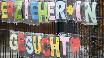 Mit bunten Buchstaben sind die Worte „Erzieher*in gesucht“ geformt, die an einem Zaun einer Kindertagesstätte hängen.