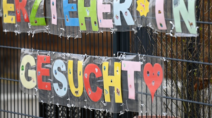 Mit bunten Buchstaben sind die Worte Erzieher*in gesucht· geformt, die an einem Zaun einer Kindertagesstätte befestigt sind.&nbsp;