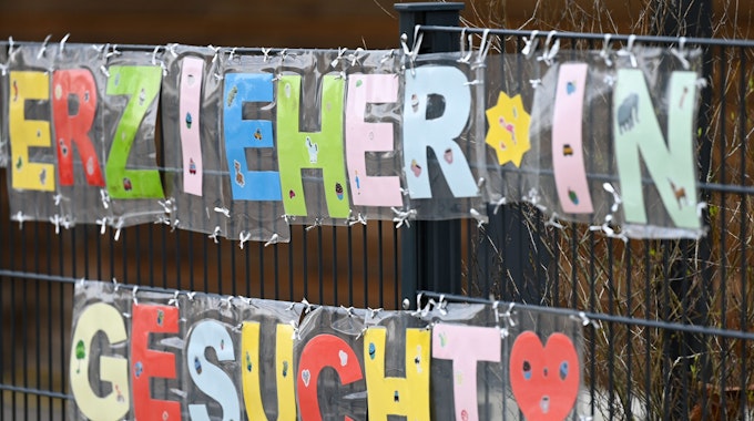 Mit bunten Buchstaben sind die Worte „Erzieher*in gesucht“ geformt, die an einem Zaun einer Kindertagesstätte hängen.