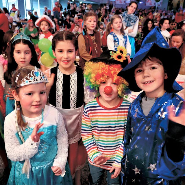 RoteSechs Kinder in Kostümen - Clowns, Prinzessinen, Einhörner und Zauberer - stehen im vollbesetzten Saal vor der Kamera und lächeln.