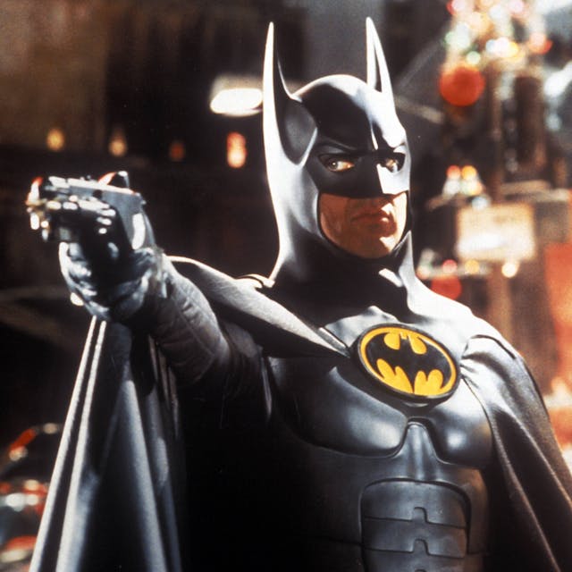 Michael Keaton als Batman, er trägt den schwarzen Batman-Anzug und hält eine Pistole in der ausgestreckten Hand.