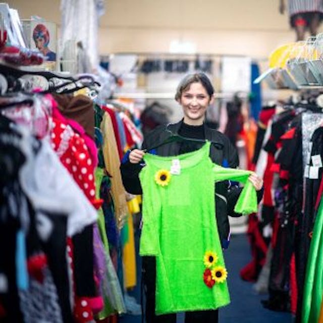 Elena Pintus hält ein grünes Kleid mit Sonnenblumen hoch. Sie steht in einem Gang zwischen Kleiderstangen mit Karnevalskostümen.