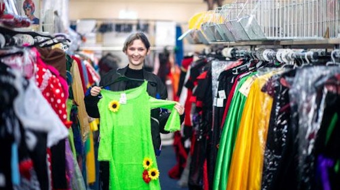 Elena Pintus hält ein grünes Kleid mit Sonnenblumen hoch. Sie steht in einem Gang zwischen Kleiderstangen mit Karnevalskostümen.