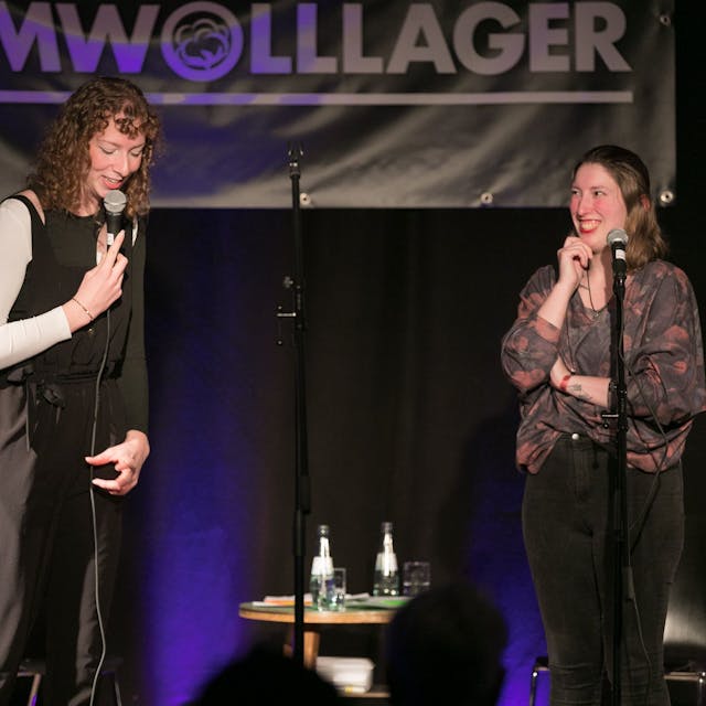 Zwei Frauen stehen auf einer Bühne: Eine spricht in ein Mikrofon, die andere schaut sie an und lacht.&nbsp;
