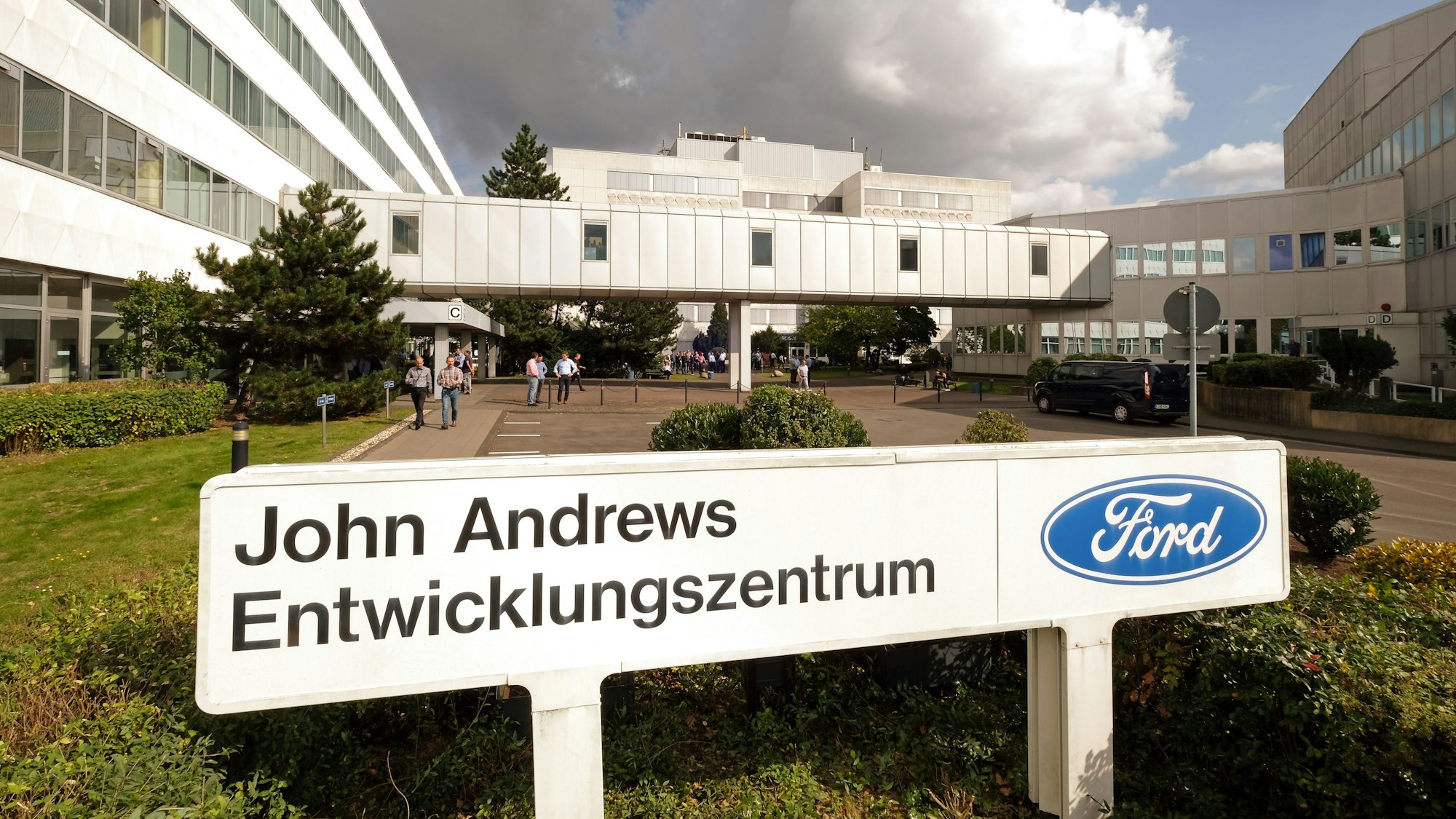 Abgebildet ist ein großes, U-förmiges Gebäude. Im Vordergrund steht ein Schild mit der Aufschrift „John Andrews Entwicklungszentrum“ mit dem blauen, ovalen Ford-Logo.