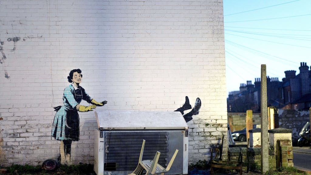 Dieses Foto zeigt eines der neuen Werke von Banksy, das eine Hausfrau aus den 1950er Jahren zu zeigen scheint, die eine klassische blaue Schürze und gelbe Abwaschhandschuhe trägt, mit einem geschwollenen Auge und einem fehlenden Zahn, die ihren männlichen Partner in eine Gefriertruhe zu schubsen scheint.&nbsp;