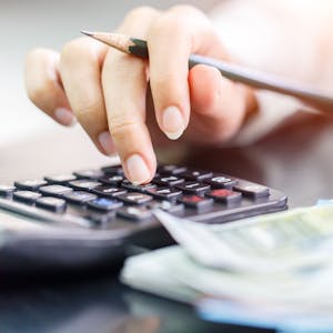 Eine Frau hält einen Bleistift in einer Hand und tippt auf einem Taschenrechner, daneben liegen Geldscheine.