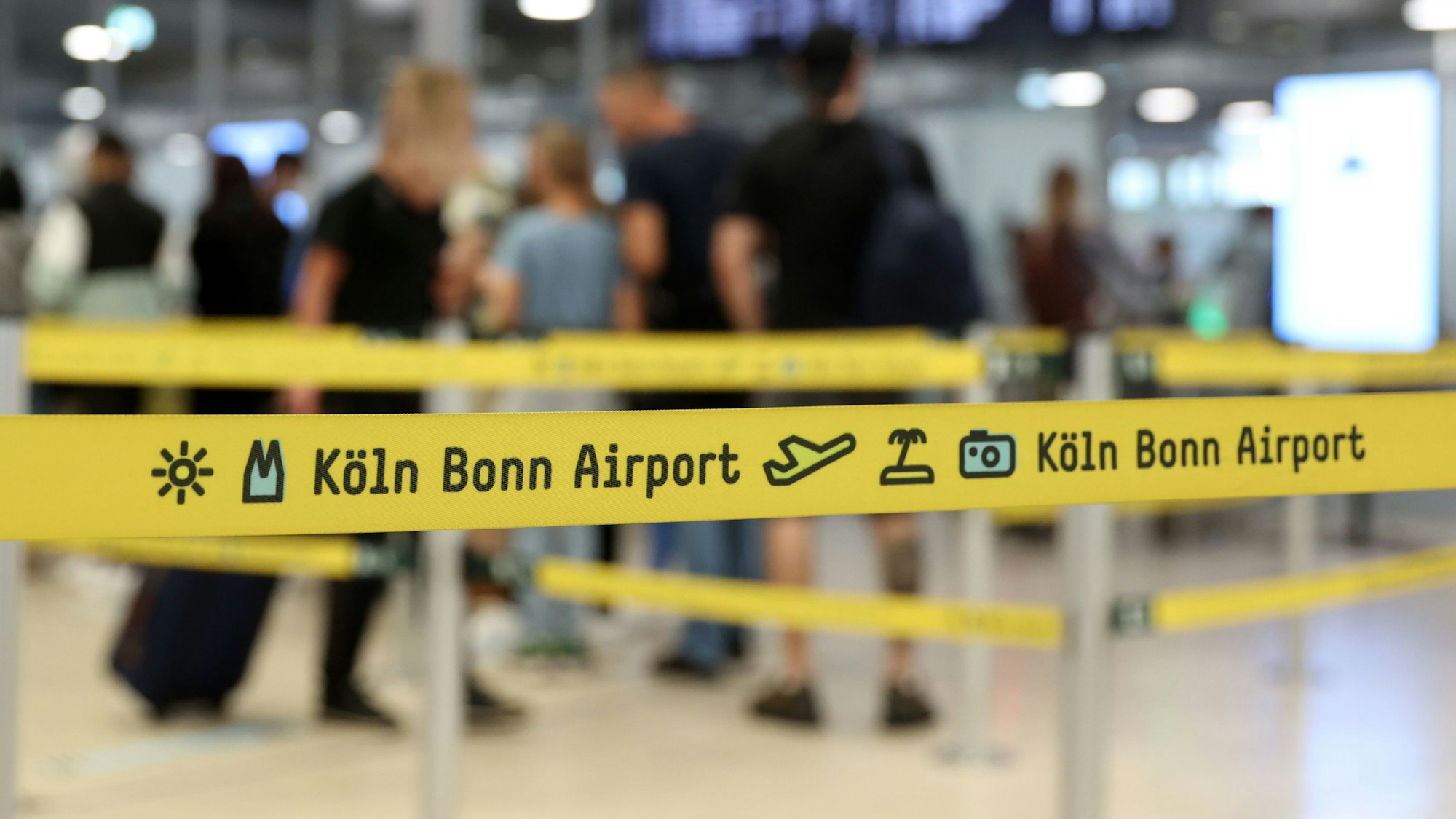 Ein gelbes Absperrband am Flughafen Köln/Bonn. Im Hintergrund sind wartende Passagiere an der Sicherheitskontrolle zu sehen.