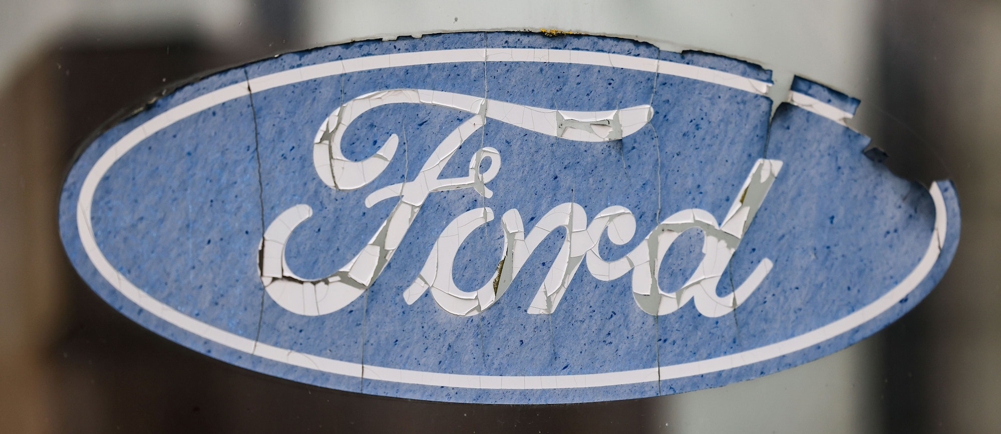 Ein abgeblättertes Logo von Ford hängt an einer Scheibe vor dem Werksgelände.