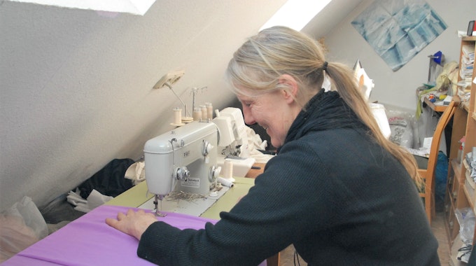 Das Foto zeigt Bekleidungsgestalterin Ursula Fischer. An ihrer Nähmaschine schneidert sie aus lila Stoff ein Kostüm.
