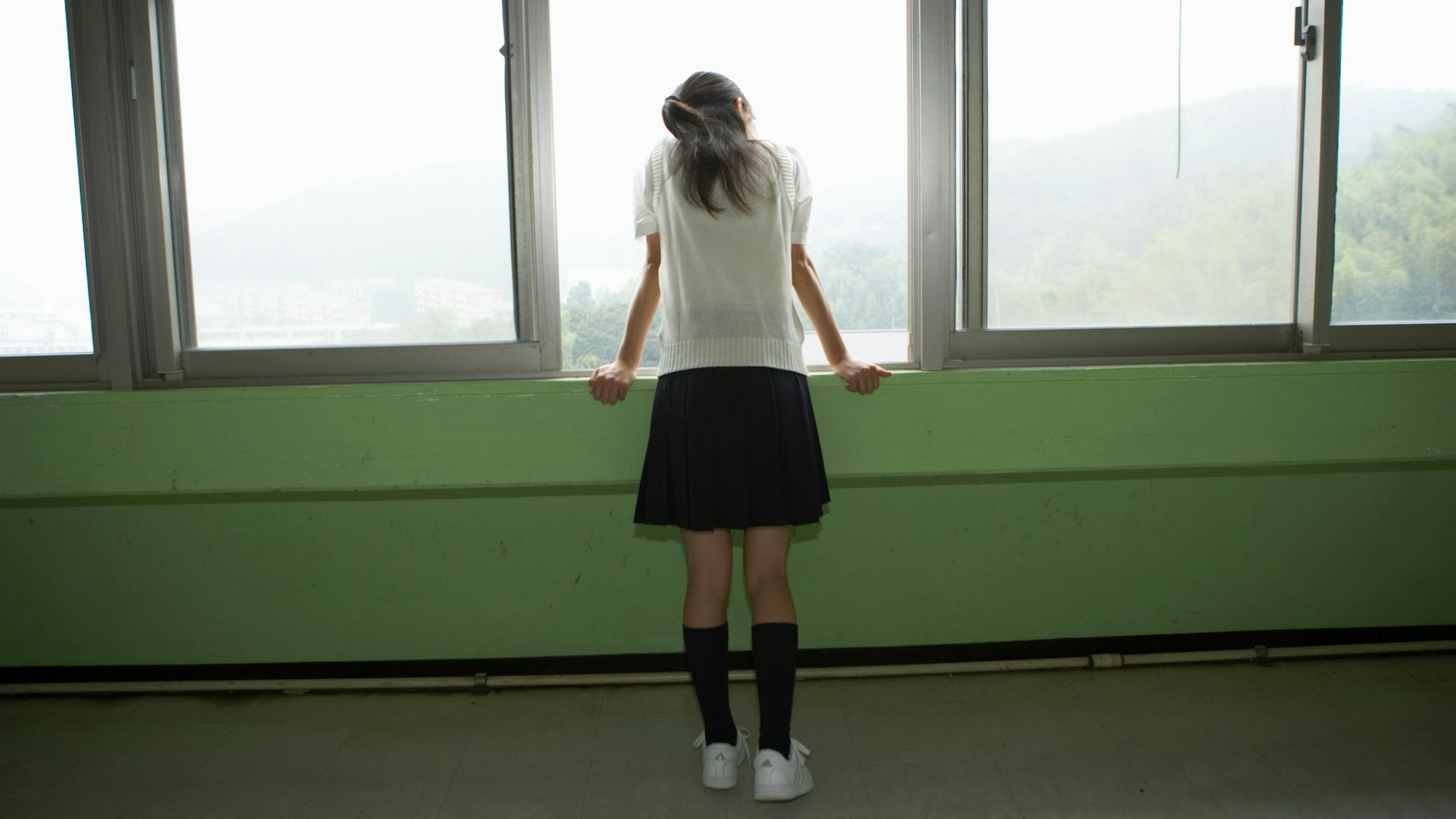 Ein Mädchen mit kurzem Rock und weißen Sneakers steht alleine an einem geöffneten Fenster.
