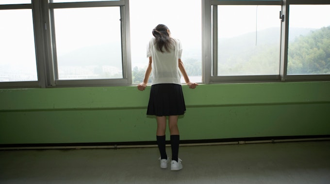 Ein Mädchen mit kurzem Rock und weißen Sneakers steht alleine an einem geöffneten Fenster.