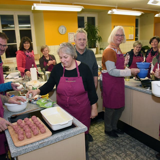 Mehrere Personen mit roten Schürzen stehen in einer großen Küche und sind Essen am Zubereiten.