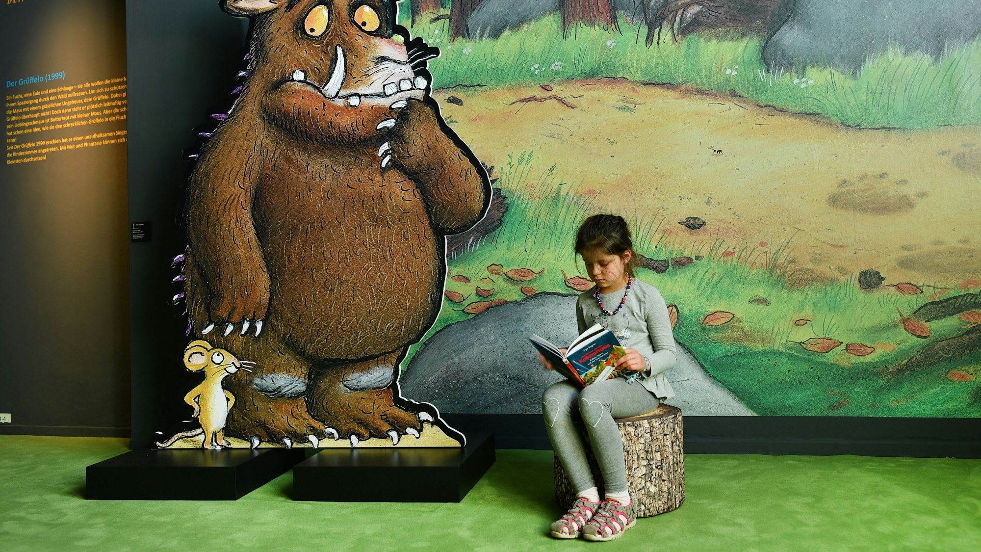 Ein Mädchen sitzt neben einer Grüffelo-Figur und liest.