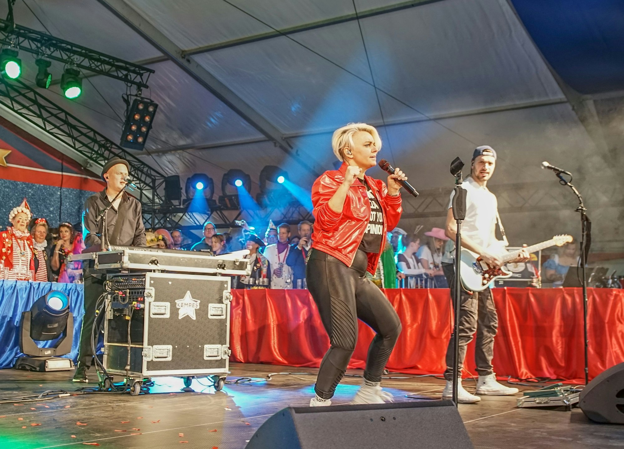 Die Mitglieder der Band stehen auf der Bühne. Sängerin Nici Kempermann trägt eine rote Lederjacke und hält das Mikrofon in der Hand.