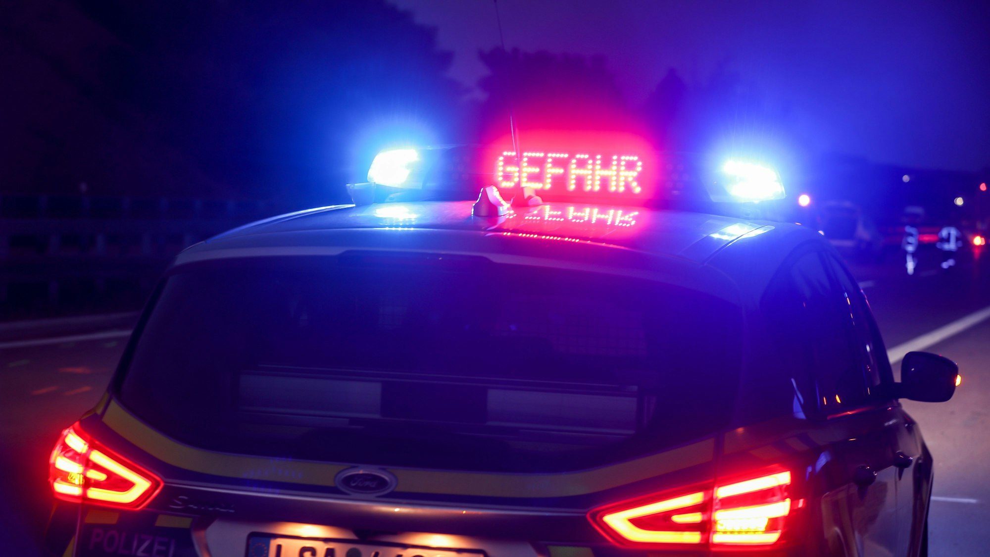 Ein Polizeiauto hat das Blaulicht an, außerdem ist in roten Buchstaben „Gefahr“ auf dem Dach des Einsatzwagens zu lesen. Im Hintergrund ist eine Unfallstelle zu sehen. (Symbolbild)