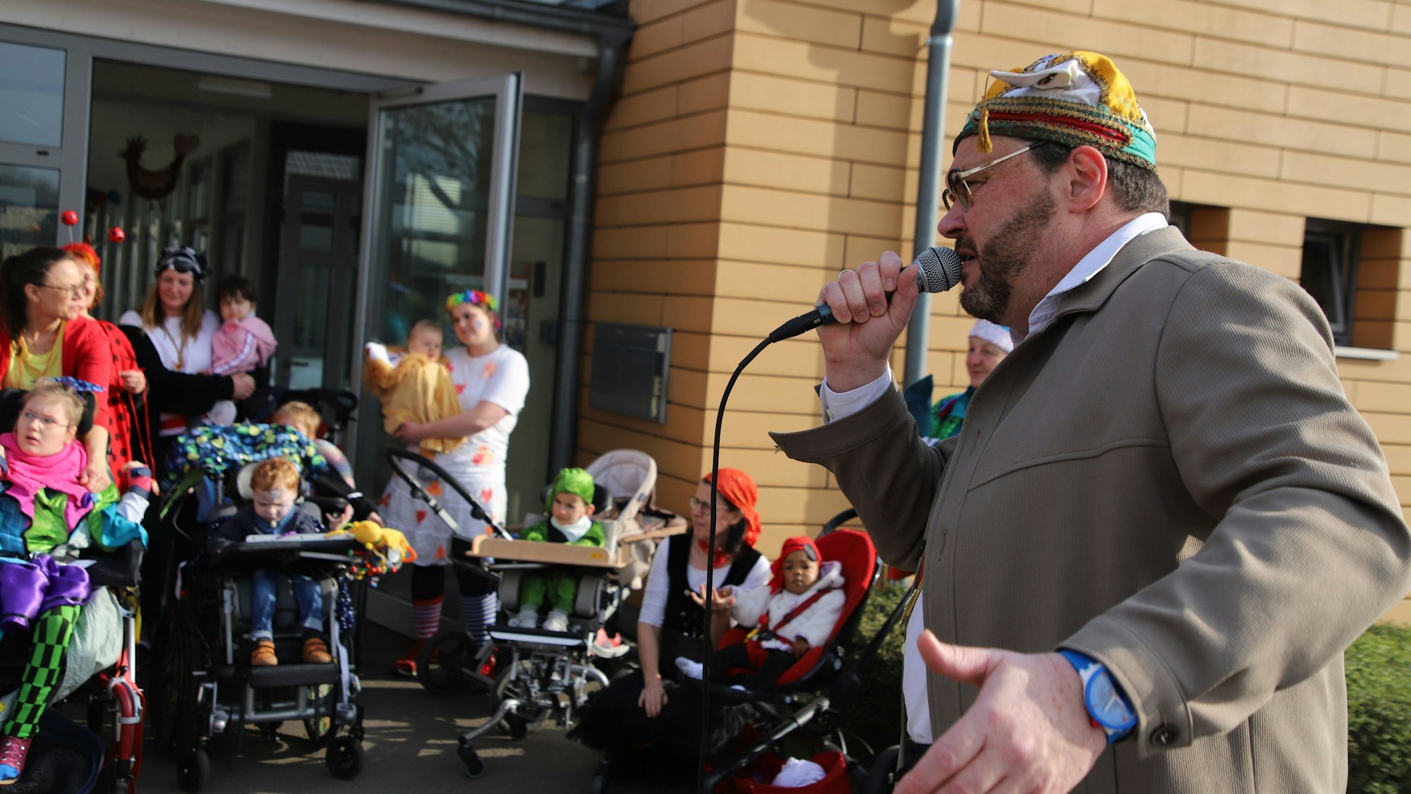 Ein Mann mit bunter Narrenkappe singt in ein Mikrofon, kostümierte Kinder in Rollstühlen und ebenfalls Frauen hören ihm vor einer Glastür eines Hauses zu.