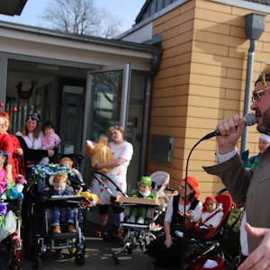 Ein Mann mit bunter Narrenkappe singt in ein Mikrofon, kostümierte Kinder in Rollstühlen und&nbsp; &nbsp;ebenfalls Frauen hören ihm vor einer Glastür eines Hauses zu.