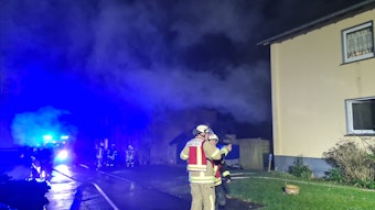 Feuerwehrleute stehen nachts im Scheinwerferlicht vor einem Wohnhaus, aus dem Brandrauch quillt.
