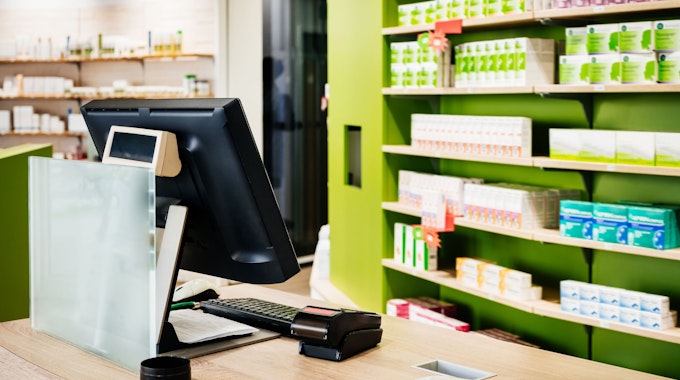Das Bild zeigt eine Apotheke mit Kasse und einem Regal mit Medikamenten.