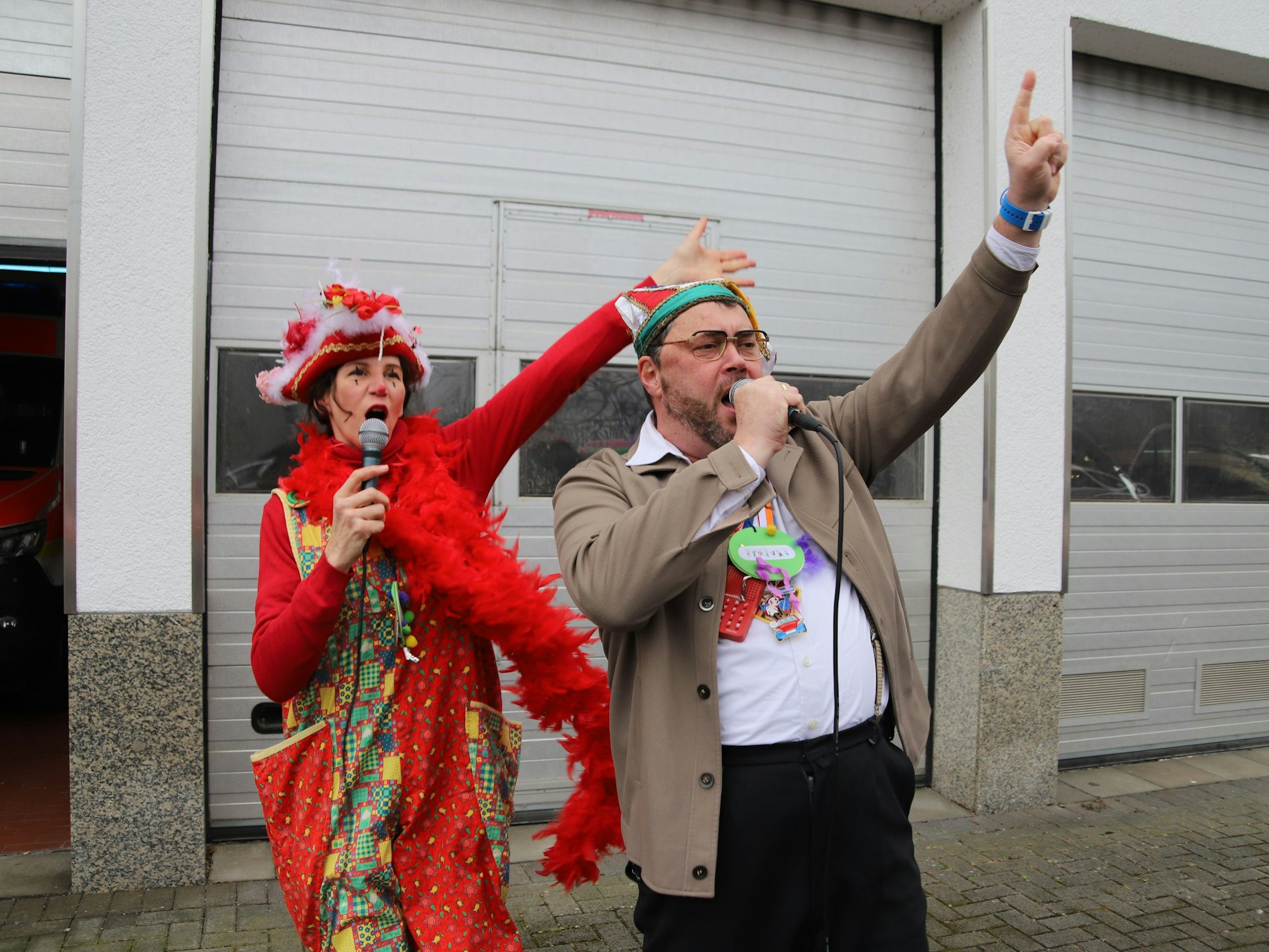 Eine Frau in Clownshose mit Hut und Federboa und ein Mann mit grauer Jacke und Narrenkappe singen in Mikrofone, sie haben den rechten Arm in die Luft gestreckt.