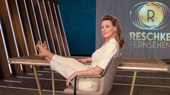 Anja Reschke sitzt auf einem Stuhl, ist zurückgelehnt und hat die Beine auf einem Tisch überschlagen. Im Hintergrund das Studio und die Aufschrift "Reschke Fernsehen"