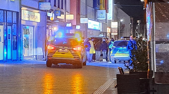 Zwei Streifenwagen stehen mit Blaulicht in der Bergisch Gladbacher Fußgängerzone. Polizeibeamte stehen bei einer Ansammlung von Menschen.