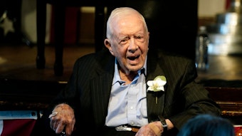 Ex-US-Präsident Jimmy Carter sitzt auf einem Stuhl und lächelt in die Kamera. Er trägt einen Anzug, in dessen Knopfloch eine Blume gesteckt ist.