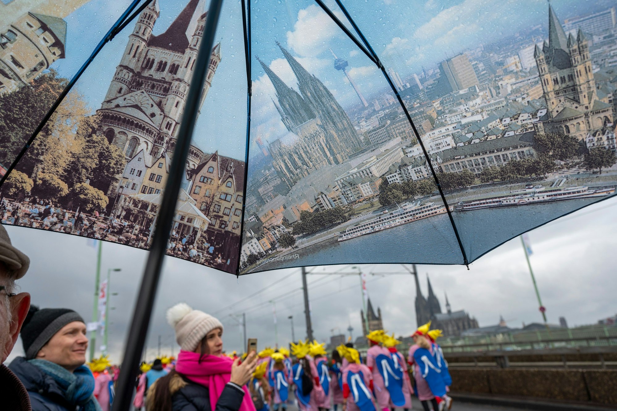 Karnevalisten stehen auf der Deutzer Brücke unter einem Regenschirm, der verschiedenen Stadtansichten Kölns zeigt.