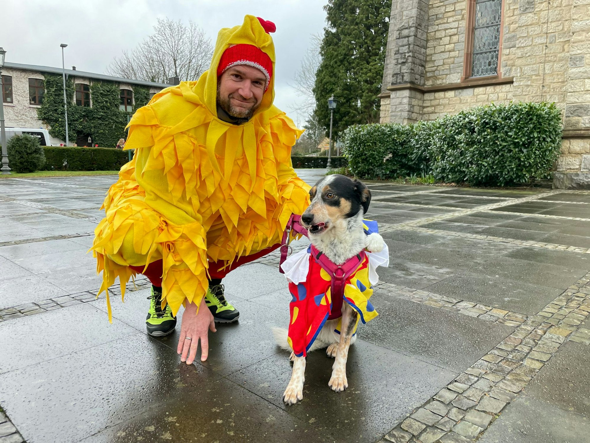 Ein Mann kniet als Huhn verkleidet neben seinem Hund, der ein Clownskostüm trägt.