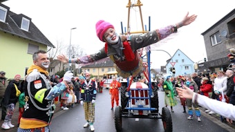 Eine bunte kostümierte Clownin schwebt als fliegender Clown an einer speziellen Konstruktion eines Traktors. Am Straßenrand stehen viele ebenfalls kostümierte Zugbesucher.