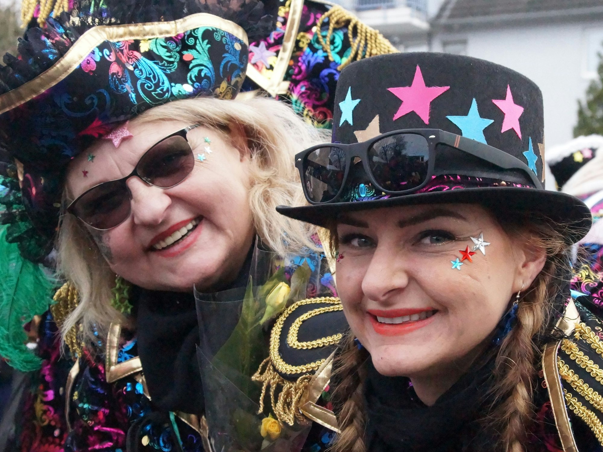 Zwei Frauen mit schwarz-bunten Hüten und Jacken lächeln in die Kamera.
