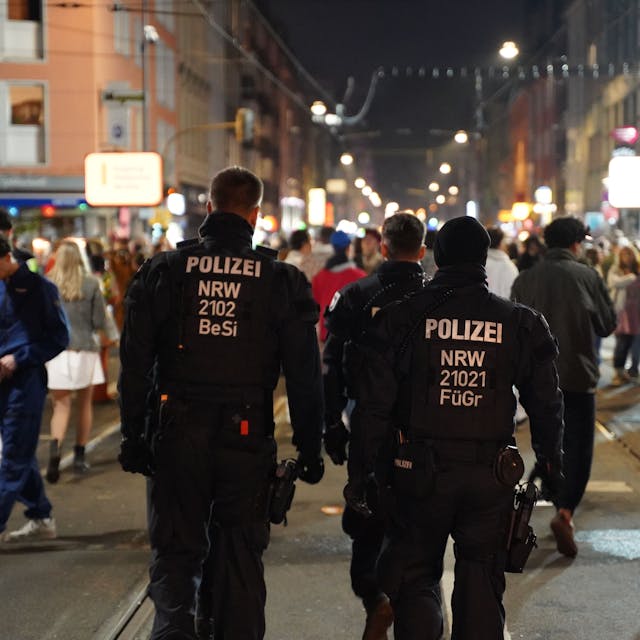 Zwei Polizisten gehen in Uniform über die Zülpicher Straße. Im Hintergrund sind zahlreiche Menschen zu erkennen, die Karneval feiern.