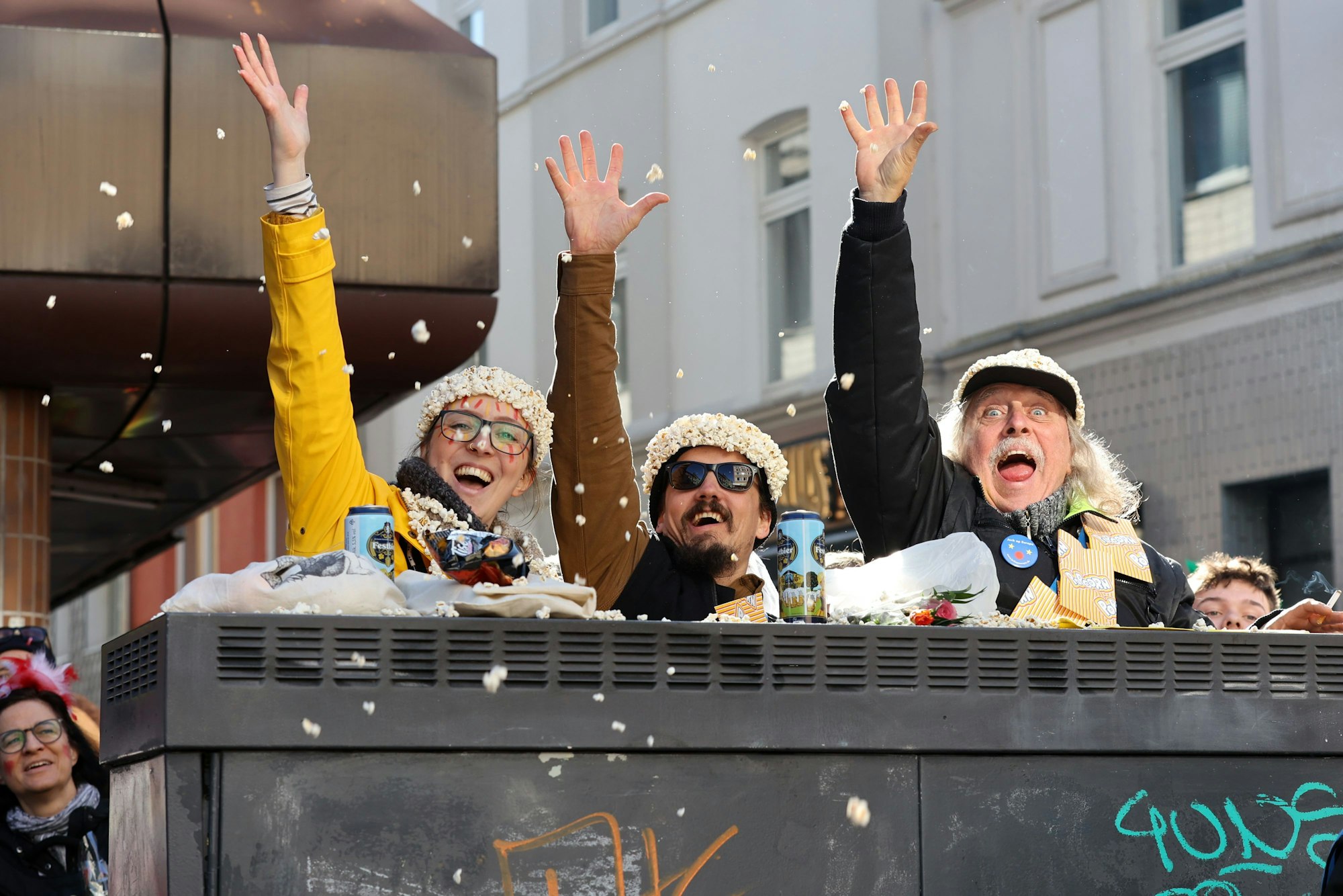 Dreie kostümierte Zuschauende tragen Kopfbedeckungen aus Popcorn und blicken lachend in die Kamera.